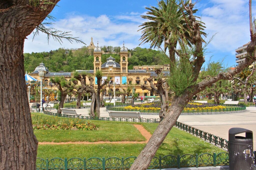 Blick durch mediterrane Bäume und Palmen auf das prachtvolle, sandsteinfarbene Rathaus von San Sebastián im Stil der Belle Époque.