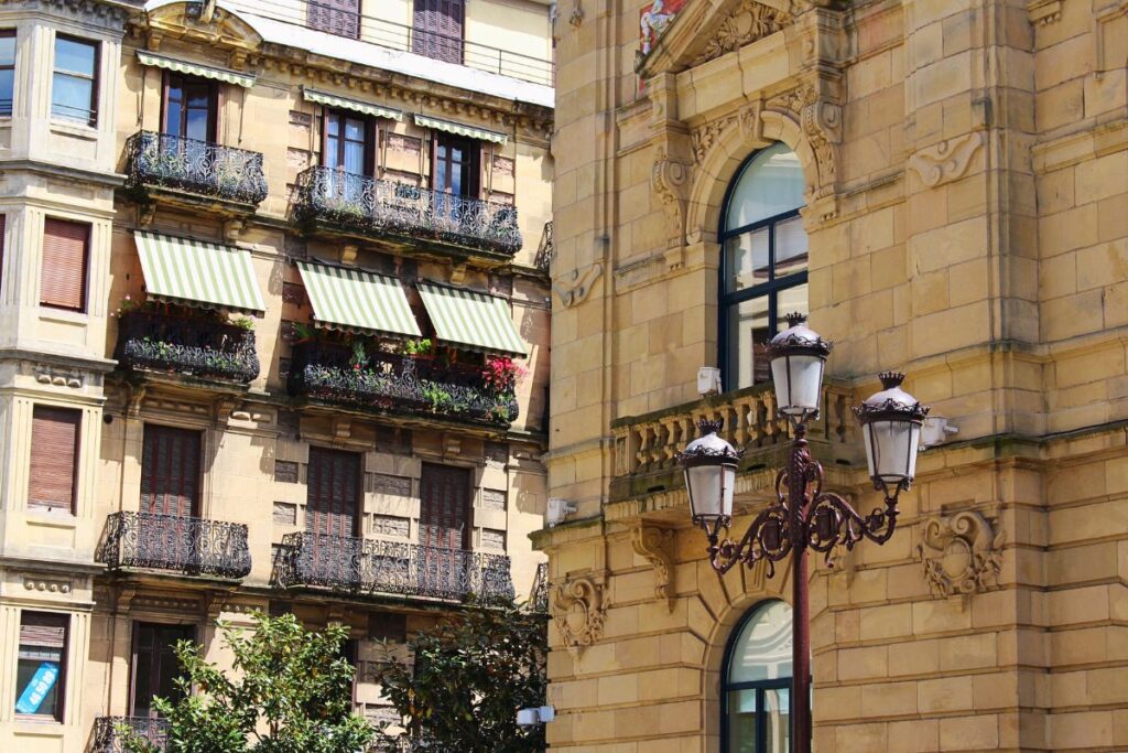 Detailansicht einer sandsteinfarbenen Palastfassade in San Sebastián mit verzierten schmiedeeisernen Balkonen, grün-weiß gestreiften Markisen und einer historischen Straßenlaterne.