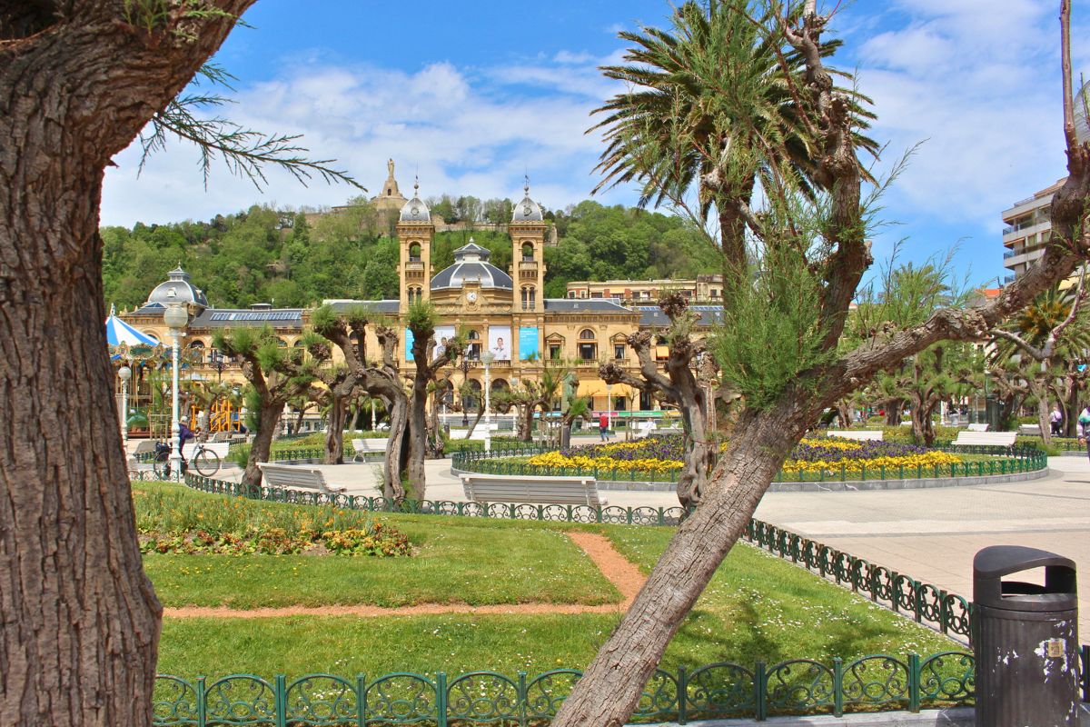 Blick durch mediterrane Bäume und Palmen auf das prachtvolle, sandsteinfarbene Rathaus von San Sebastián im Stil der Belle Époque.