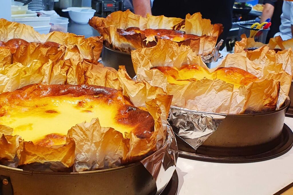 Mehrere frisch gebackene, traditionelle baskische Käsekuchen (Tarta de Queso) in runden Backformen mit Pergamentpapier auf dem Tresen der Bar La Viña in San Sebastián.