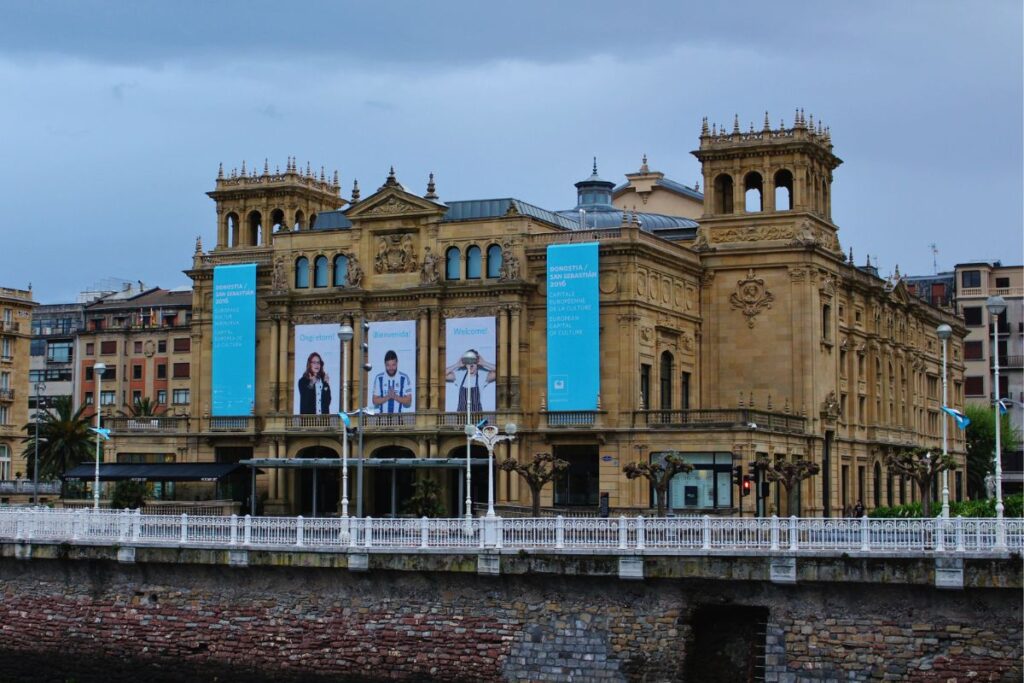Die herrschaftliche Fassade des Victoria Eugenia Theaters in San Sebastián aus hellem Sandstein mit großen blauen Veranstaltungsbannern am Ufer des Flusses Urumea.
