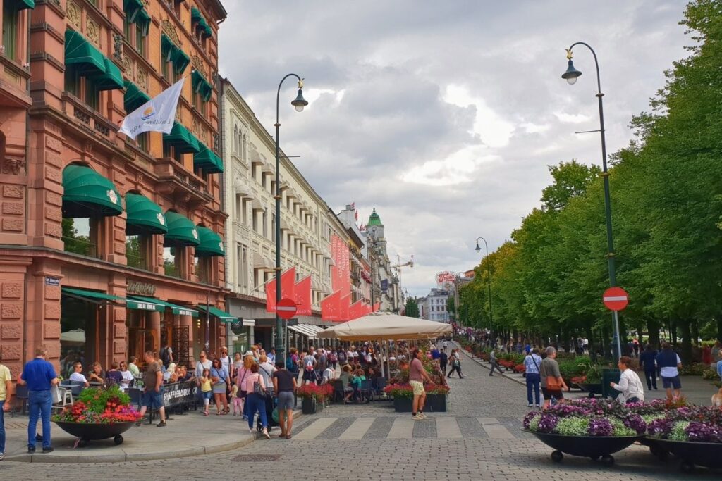 Die belebte Einkaufsstraße Karl Johans gate in Oslo, gesäumt von Bäumen, historischen Gebäuden wie dem Grand Hotel und vollen Straßencafés.