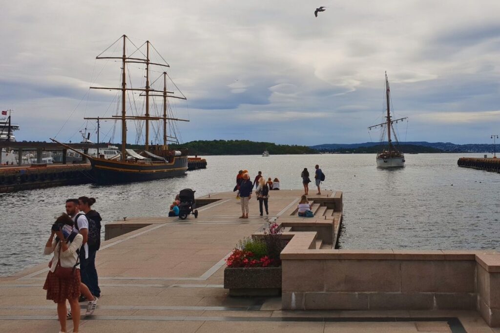 Touristen spazieren auf dem steinernen Pier am Osloer Hafen, im Hintergrund liegen traditionelle Segelschiffe vor Anker mit Blick auf den Oslofjord.