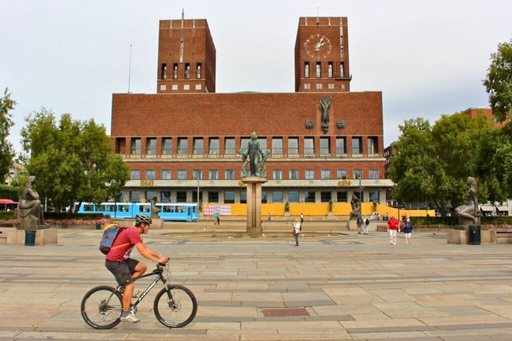 Das monumentale rote Backsteingebäude des Osloer Rathauses mit zwei Türmen, davor ein weiter Platz mit Skulpturen und einem Radfahrer im Vordergrund.