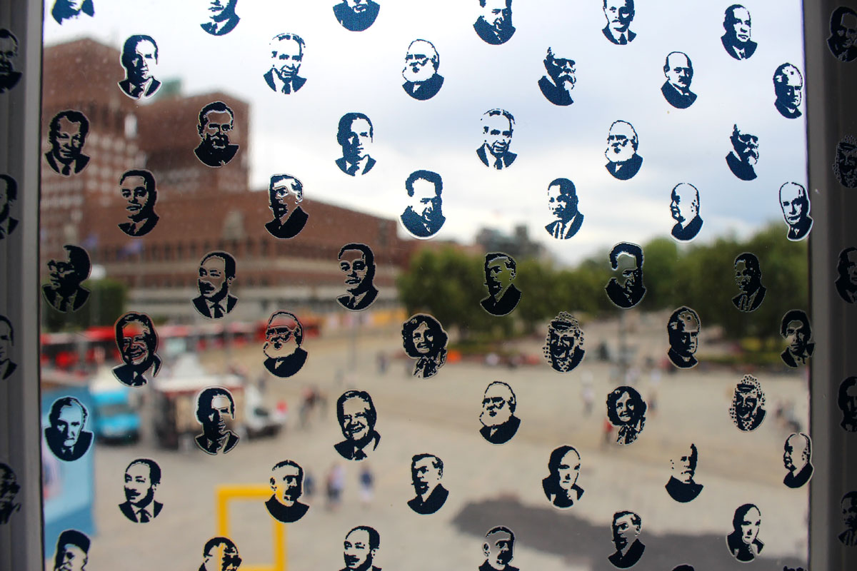 Blick durch eine Fensterscheibe, die mit kleinen Stickern von Köpfen (Nobelpreisträger) beklebt ist, auf den belebten Platz vor dem Museum.