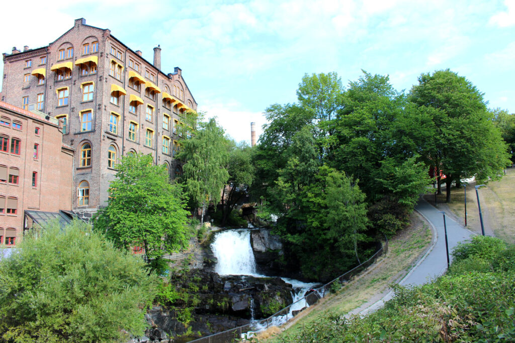 Der mächtige Mølla-Wasserfall im Fluss Akerselva in Oslo. Links steht ein hohes historisches Backsteingebäude (ehemals Fabrik) mit gelben Markisen, rechts führt ein gepflasterter Spazierweg durch grüne Bäume.
