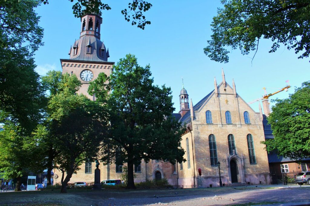 Die Osloer Domkirche mit ihrem massiven Turm und der Uhr ragt in den blauen Himmel, umgeben von großen grünen Bäumen und einem gepflasterten Platz.