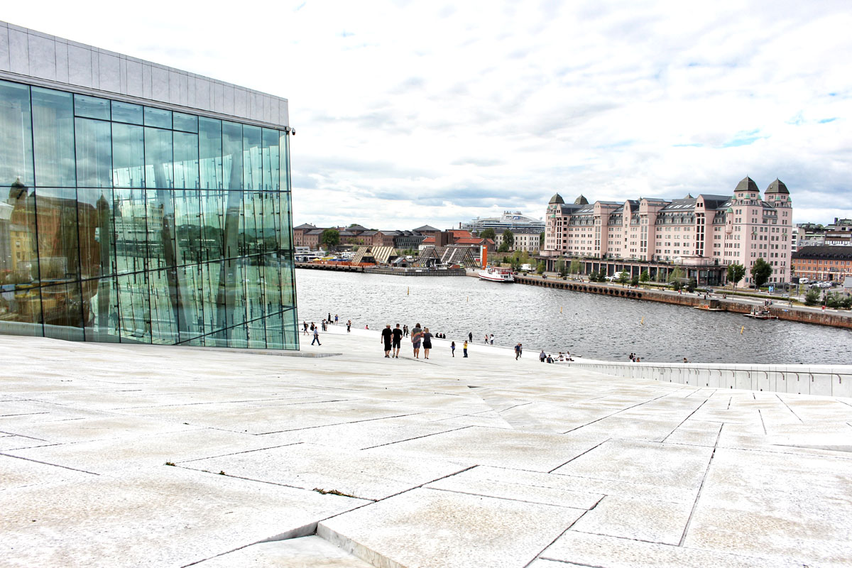 Blick vom Dach des Opernhauses über das Wasser des Oslofjords auf das rosafarbene historische Gebäude "Havnelageret", links eine große Glaswand der Oper.