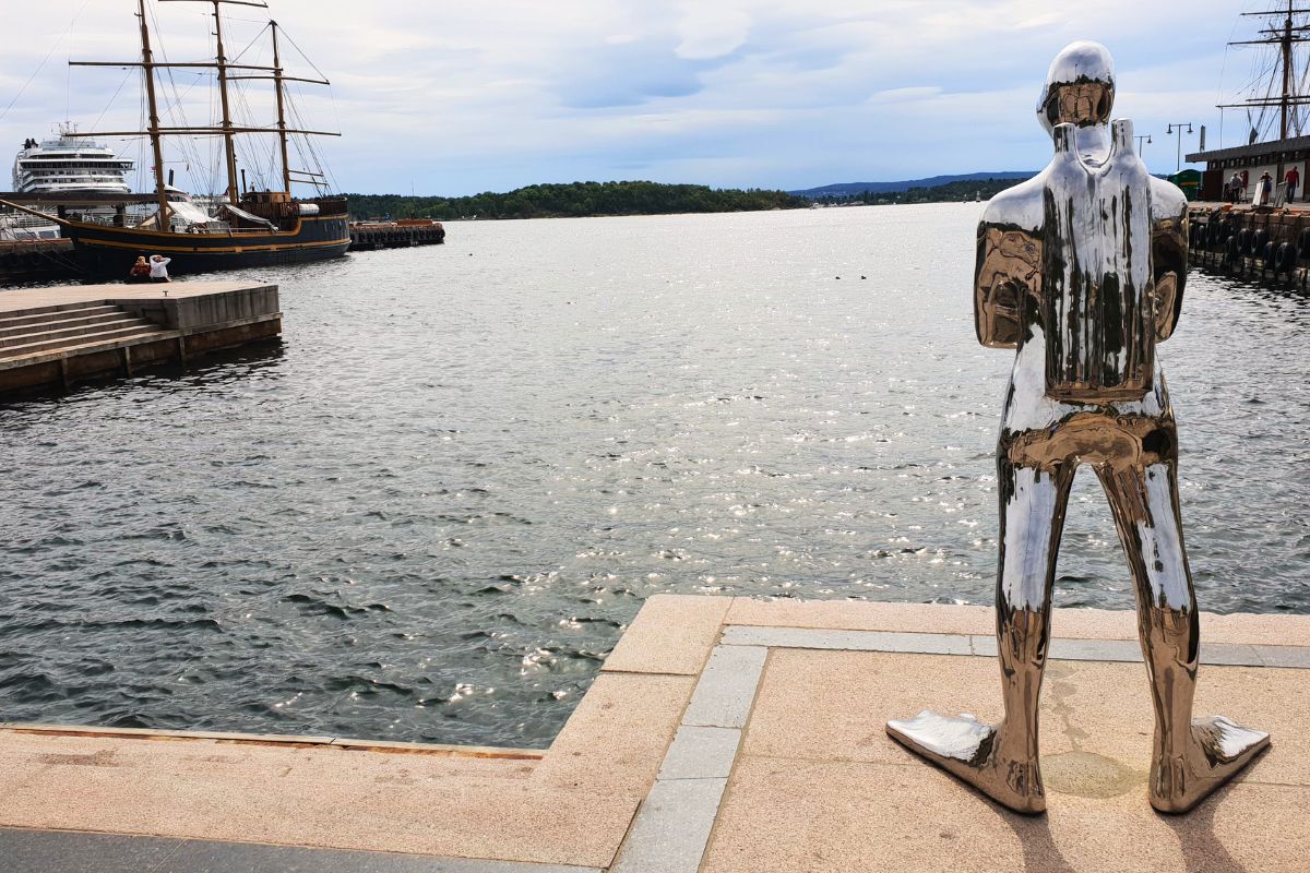 Die verchromte Skulptur eines Tauchers ("Dykkaren") steht an der Hafenpromenade und blickt hinaus auf den Oslofjord. Im Hintergrund liegen Segelschiffe und eine Fähre.