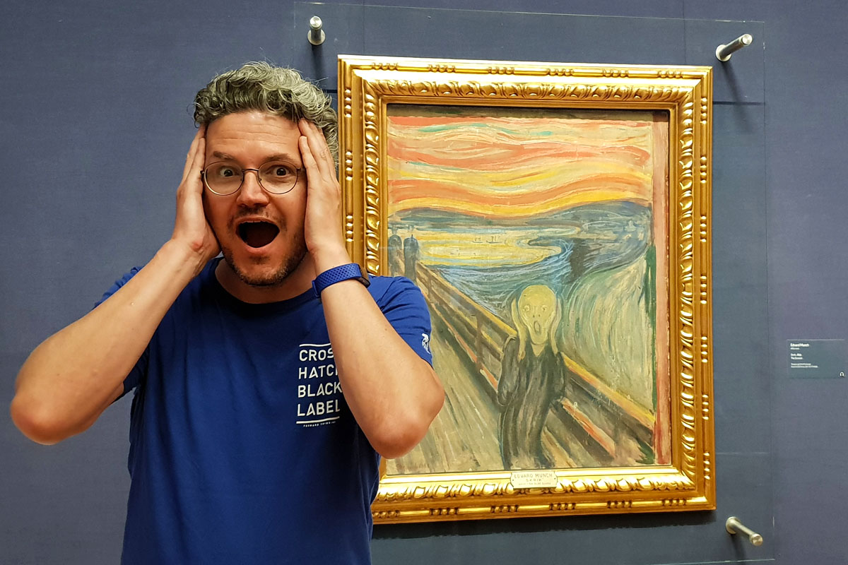 Reiseblogger Nicolo Martin steht im Nationalmuseum Oslo vor dem Gemälde „Der Schrei“ von Edvard Munch und imitiert mit weit aufgerissenem Mund und Händen an den Wangen die berühmte Pose der Figur.