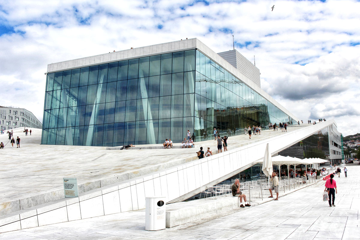 Das weiße, schräge Marmordach des Osloer Opernhauses ragt wie eine Eisscholle aus dem Wasser, im Hintergrund blauer Himmel mit Wolken. Menschen spazieren auf dem Dach.