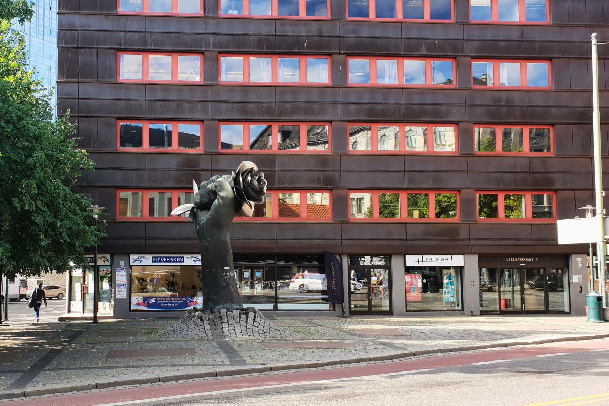 Die bronzene Skulptur "Neve med rose" (Faust mit Rose) am Lilletorget in Oslo. Eine große Hand hält eine Rose fest umschlossen, im Hintergrund ein rotes Bürogebäude.