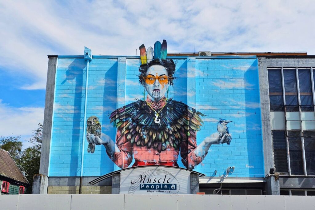 Ein riesiges, blaues Wandgemälde an einem Gebäude in Christchurch, das eine Frau in traditioneller Māori-Kleidung mit zwei Vögeln darstellt.
