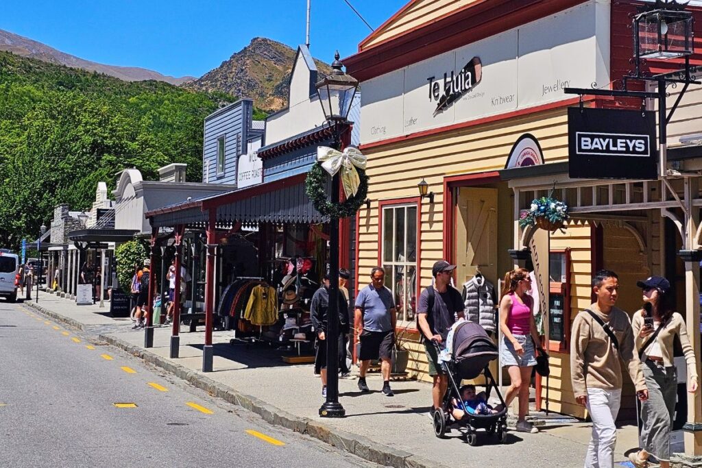 Eine belebte historische Straße in Arrowtown mit flachen, bunten Goldgräber-Häusern, Geschäften und Menschen, die bei sonnigem Wetter spazieren gehen.