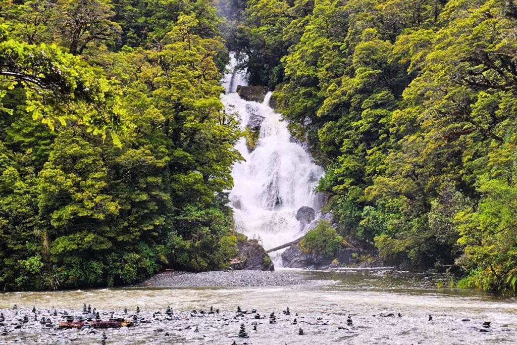 Der weiße, fächerförmige Wasserfall Fantail Falls stürzt inmitten eines grünen Waldes am Haast Pass in ein Flussbett mit vielen Steinmännchen im Vordergrund.