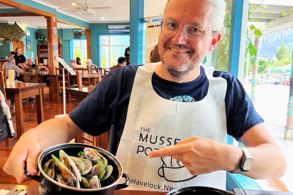 Reiseblogger Nicolo Martin zeigt lächelnd auf einen Topf mit neuseeländischen Grünlippmuscheln im Restaurant „The Mussel Pot“ in Havelock.