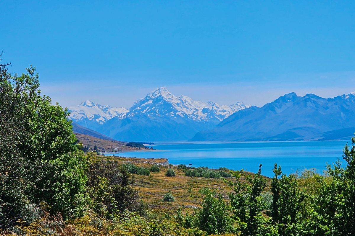 Panorama-Aussicht über den türkisblauen Lake Pukaki auf den schneebedeckten Gipfel des Mount Cook (Aoraki) unter strahlend blauem Himmel.