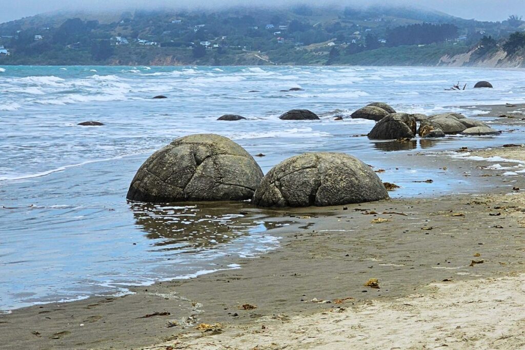 Mehrere große, kugelrunde Moeraki Boulders liegen im Sand und im seichten Wasser am Koekohe Beach.