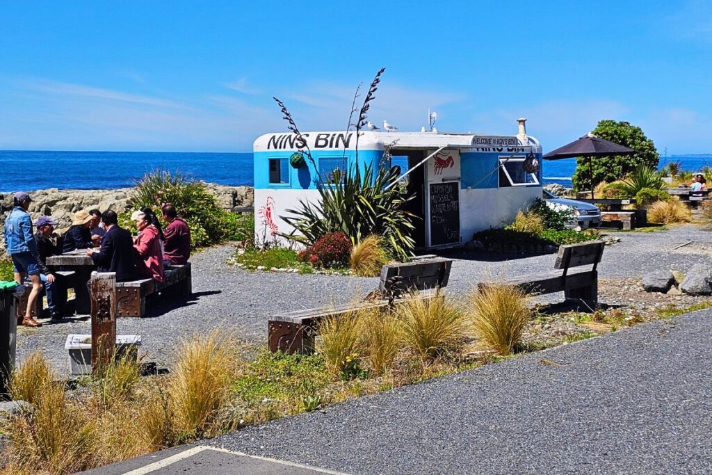 Ein blau-weißer Imbisswagen namens „Nin's Bin“ direkt an der Küste von Kaikoura, an dem Menschen an Holztischen frische Meeresfrüchte genießen.