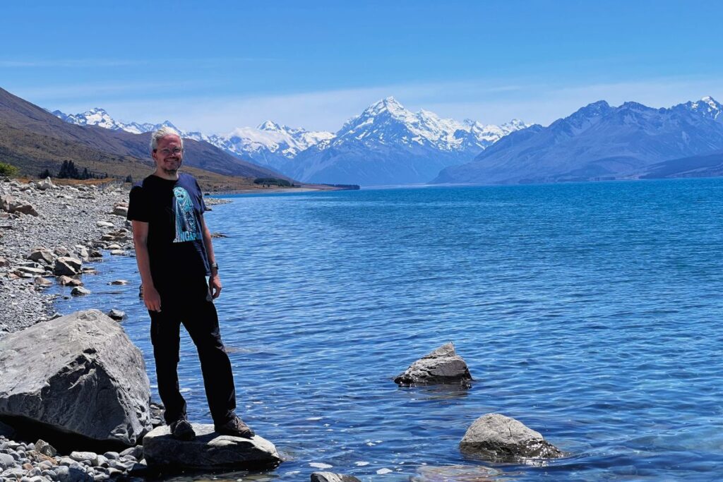 Reiseblogger Nicolo Martin steht lächelnd auf Steinen am Ufer des Lake Pukaki vor der Kulisse des Mount Cook (Aoraki).