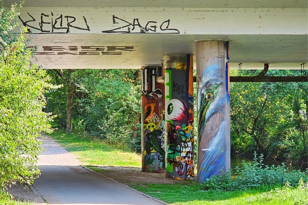 Graffiti mit bunten Motiven und Vogelauge an Brückenpfeiler entlang der Alb in Karlsruhe