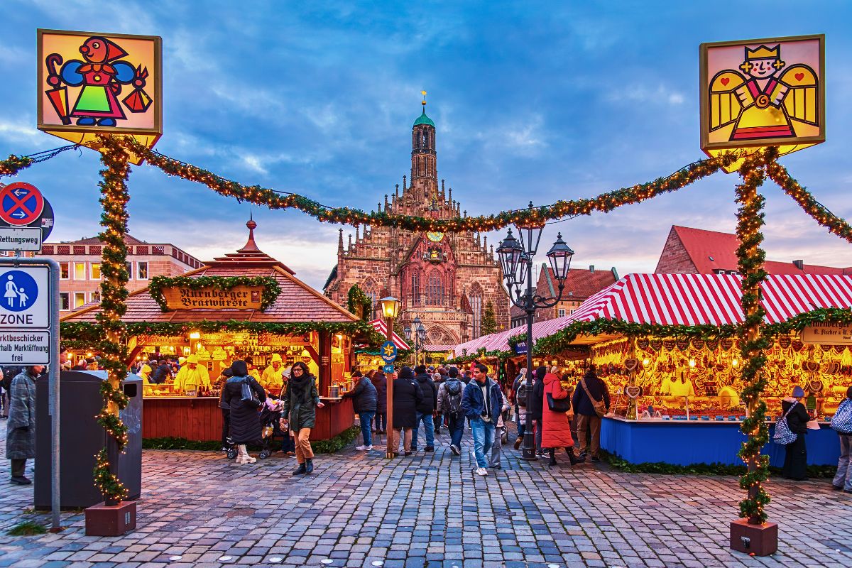 Stimmungsvolle Übersicht des Nürnberger Christkindlesmarktes zur blauen Stunde, mit der Frauenkirche im Hintergrund, beleuchteten Buden, den markanten Zwetschgenmännle-Schildern am Eingang und vielen Besuchern