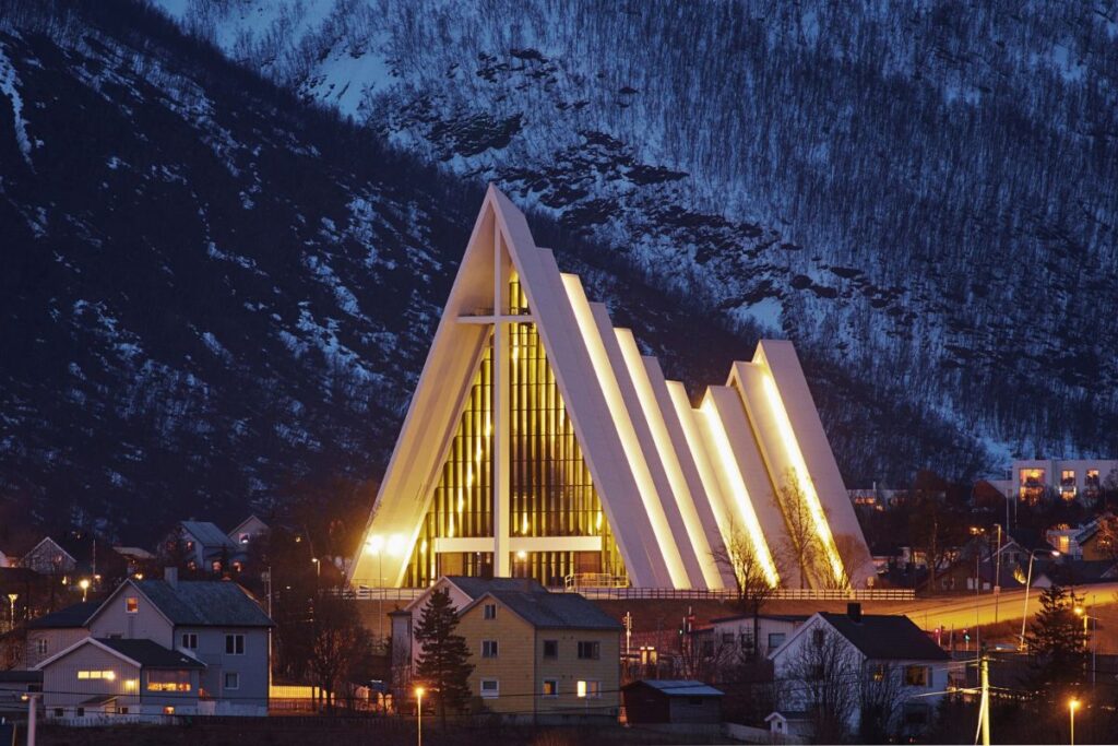 Die beleuchtete Eismeerkathedrale in Tromsø vor verschneitem Bergmassiv bei Abenddämmerung