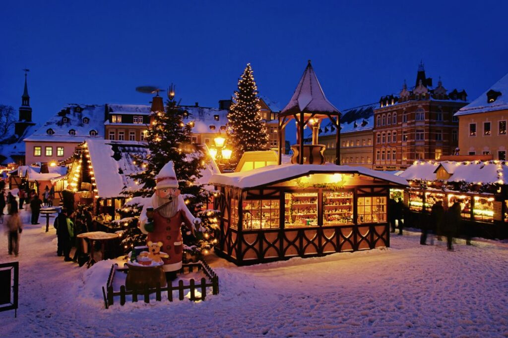 Verschneiter Weihnachtsmarkt in Annaberg-Buchholz mit großer hölzerner Wichtelfigur, Marktbuden und Weihnachtsbaum zur blauen Stunde.