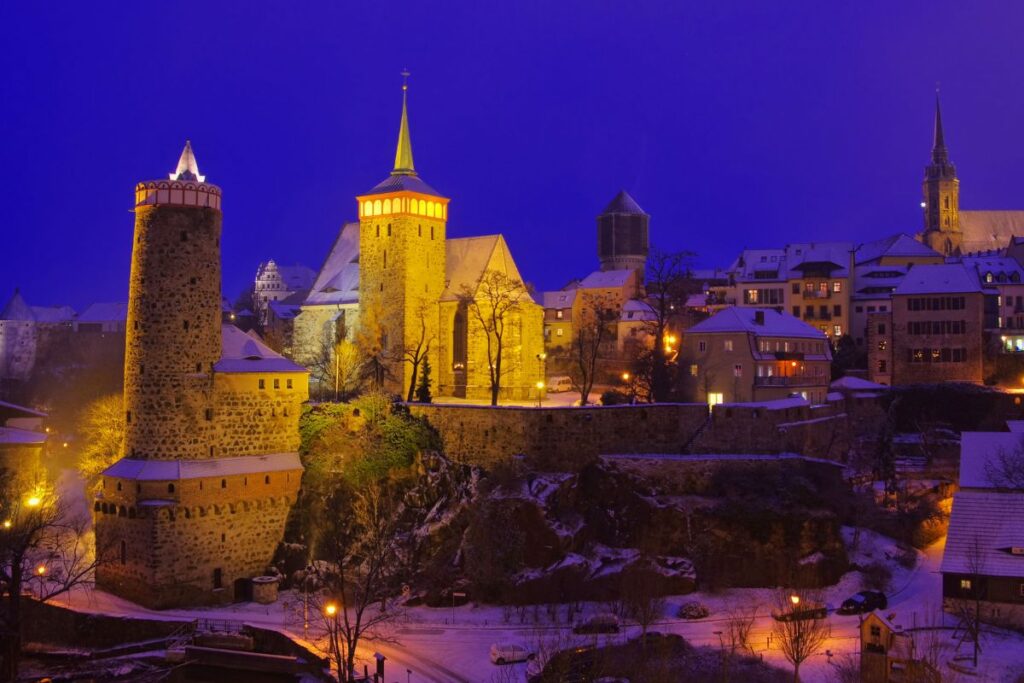 Blick auf die verschneite, blau beleuchtete Altstadt von Bautzen mit der Alten Wasserkunst und Michaeliskirche bei Nacht.