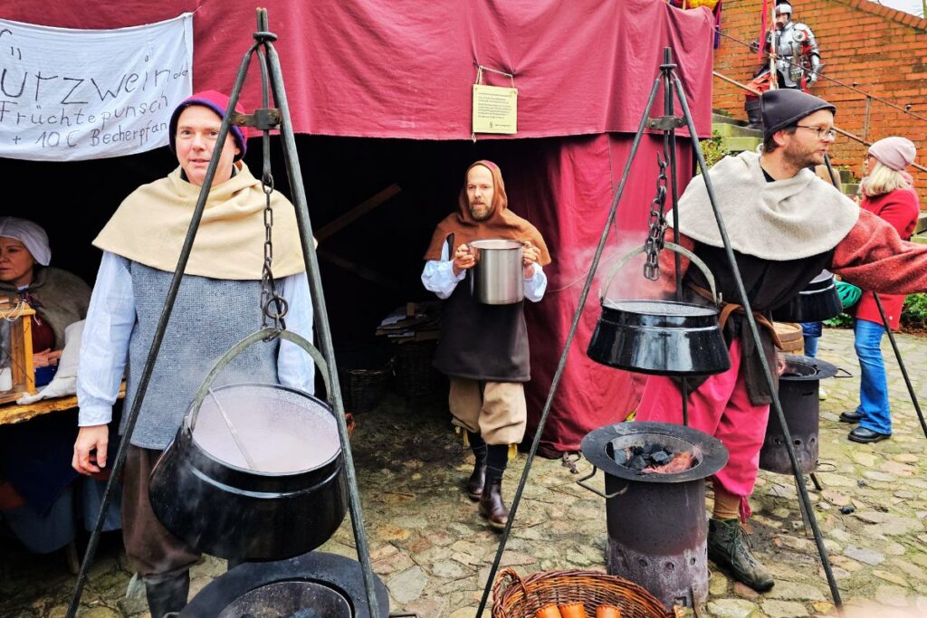 Darsteller in mittelalterlichen Gewändern kochen Glühwein und Speisen in Kesseln über offenem Feuer auf dem Historischen Christmarkt in Lüneburg.