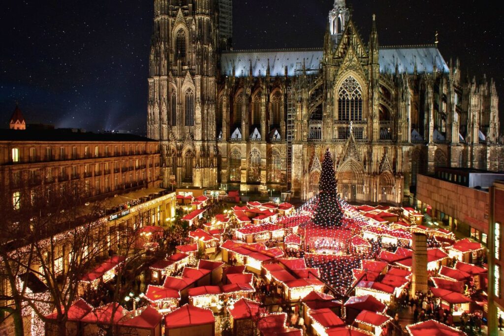 Übersicht über den Weihnachtsmarkt am Kölner Dom bei Nacht mit dem riesigen Weihnachtsbaum, den roten Dächern der Buden und dem beleuchteten Dom im Hintergrund.