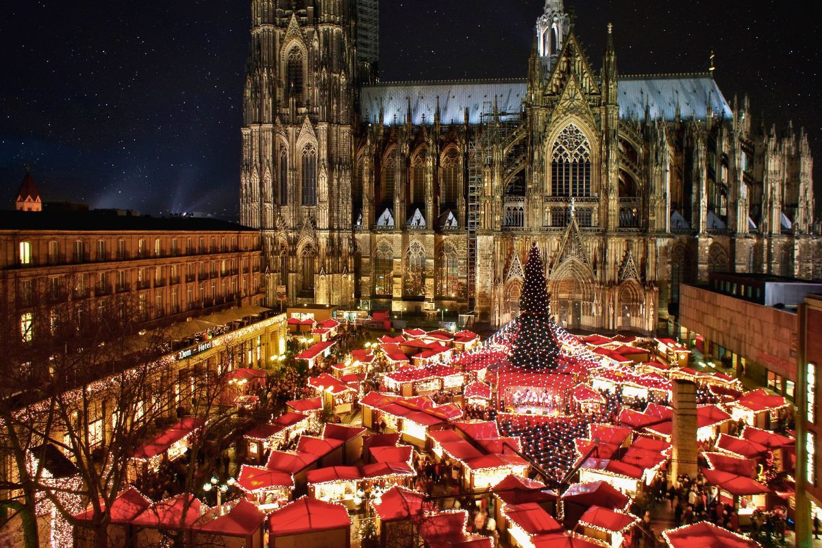 Übersicht über den Weihnachtsmarkt am Kölner Dom bei Nacht mit dem riesigen Weihnachtsbaum, den roten Dächern der Buden und dem beleuchteten Dom im Hintergrund.