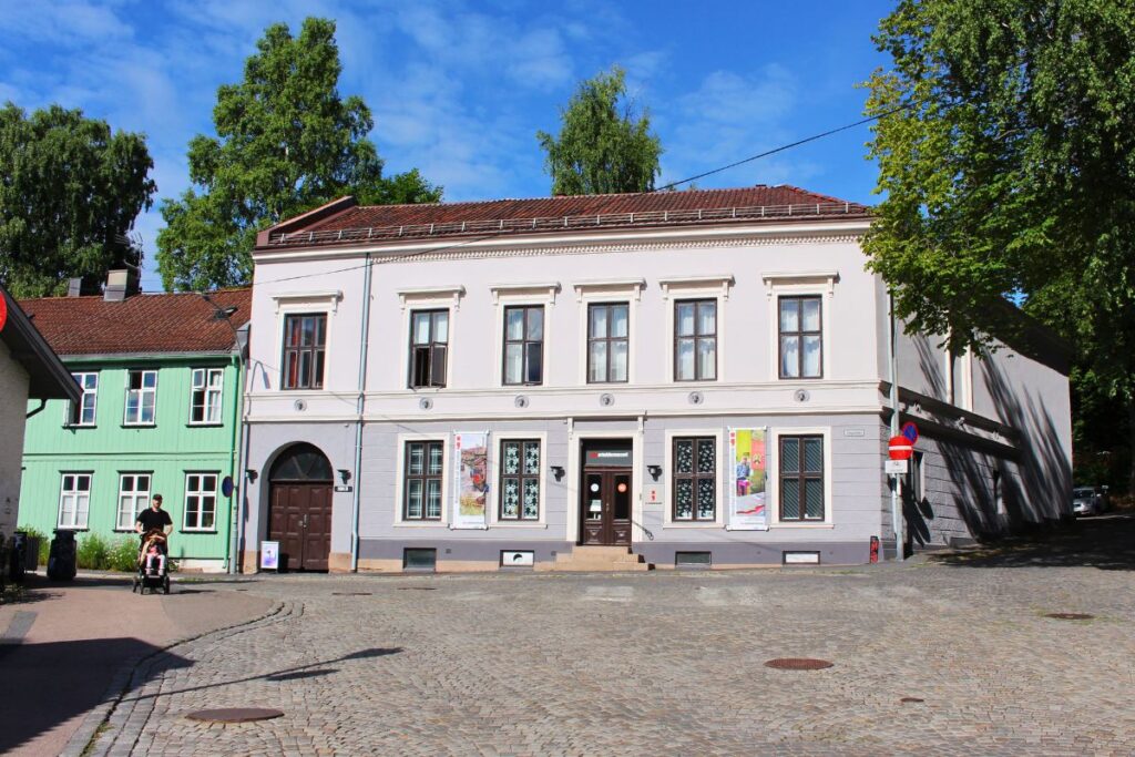Das graue Gebäude des Arbeidermuseet (Arbeitermuseum) in Oslo Sagene an einer Kopfsteinpflasterstraße neben einem historischen grünen Holzhaus