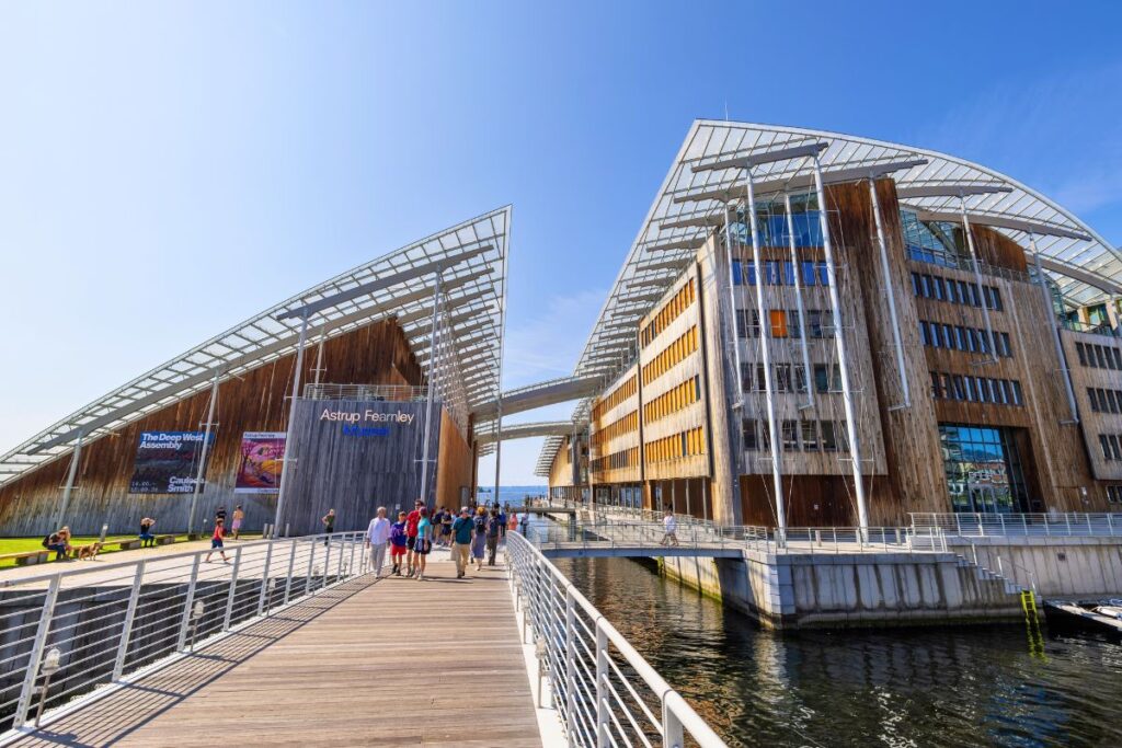 Das Astrup Fearnley Museum für Moderne Kunst in Oslo mit seinem markanten Glasdach und Holzfassade am Wasser, davor Fußgänger auf einer Brücke