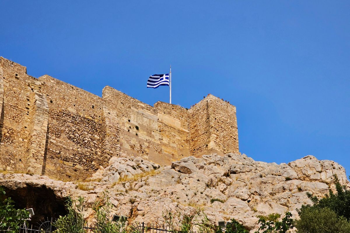 Blick von unten auf die massiven antiken Steinmauern der Akropolis in Athen, über denen die blau-weiße griechische Flagge an einem hohen Mast im Wind weht.