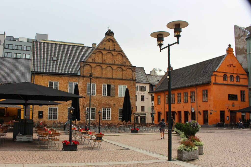 Der gepflasterte Christiania Torv Platz in Oslo mit historischen ockerfarbenen Gebäuden, dem Gamle Rådhus und Außengastronomie