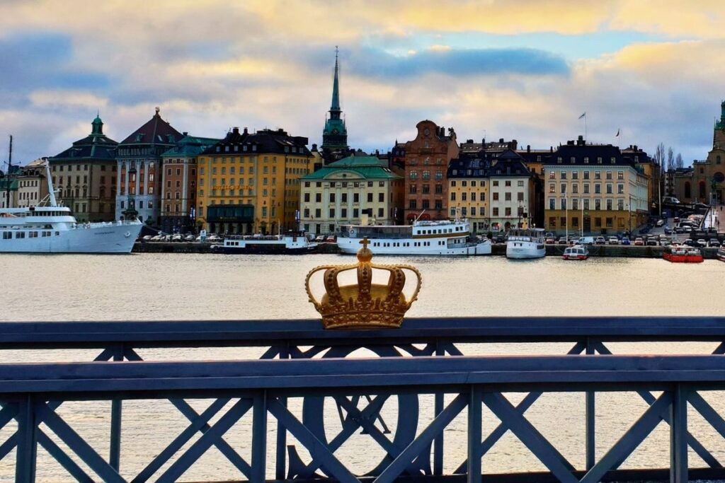 Eine goldene Krone auf dem Geländer der Skeppsholmsbron-Brücke im Vordergrund, unscharf im Hintergrund die bunten Häuser der Altstadt Gamla Stan in Stockholm.