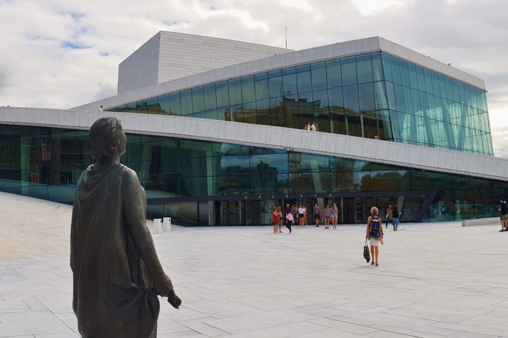 Blick über den Vorplatz der Oper Oslo auf das moderne Glasgebäude. Im Vordergrund steht die dunkle Bronzestatue der Opernsängerin Kirsten Flagstad, die zum Gebäude blickt.