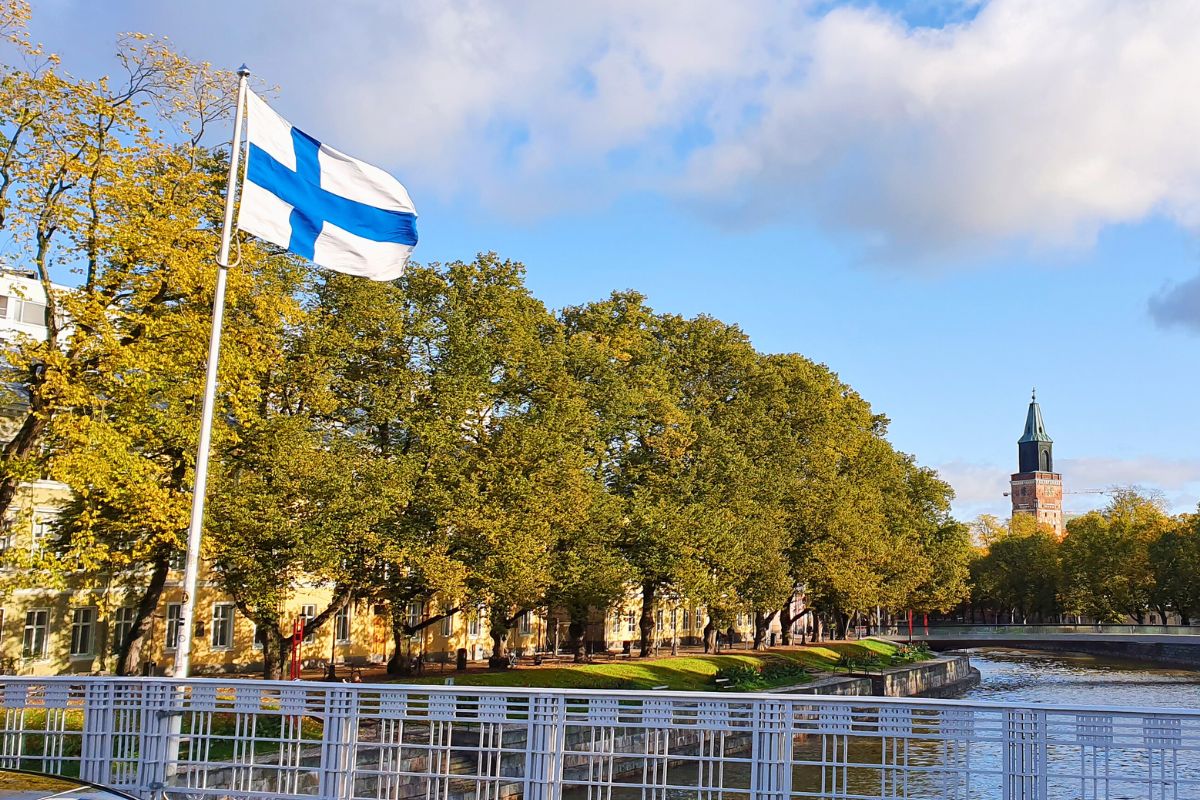 Die finnische Flagge weht im Wind vor einer sonnigen Stadtkulisse am Fluss in Turku.
