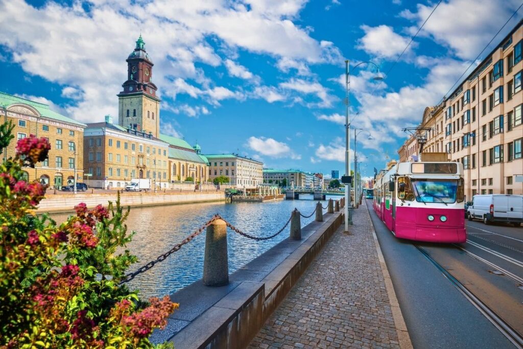 Eine typische rosa-weiße Straßenbahn fährt in Göteborg an einem Kanal entlang, im Hintergrund historische Gebäude und ein Kirchturm unter blauem Himmel.