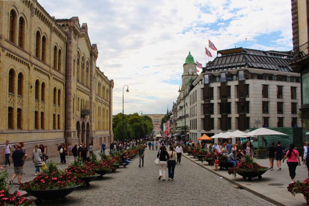 Blick auf die belebte Karl Johans gate in Oslo mit Fußgängern, Blumenkübeln und historischen Gebäuden unter bewölktem Himmel