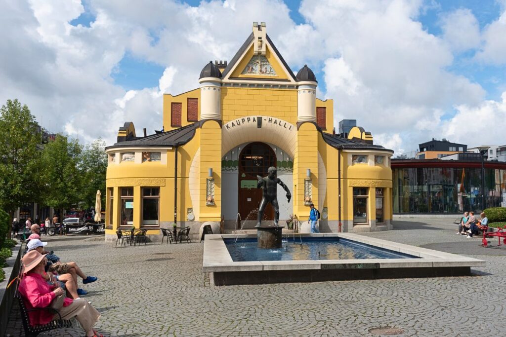 Die historische gelbe Markthalle (Kauppahalli) in Kuopio mit einem Springbrunnen und einer Bronzestatue im Vordergrund.