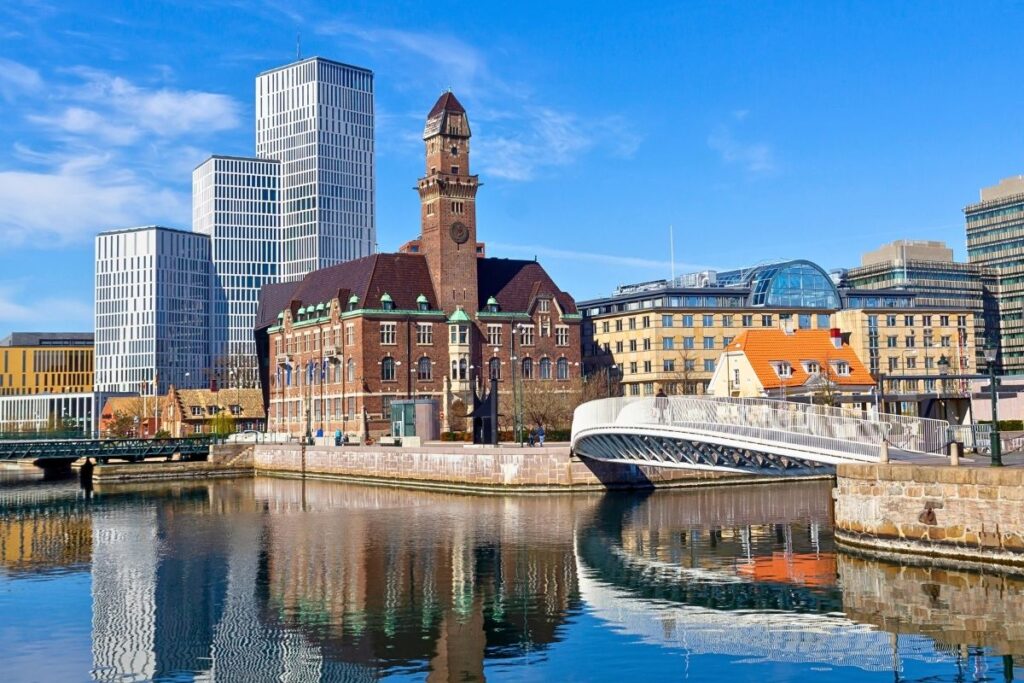 Blick über einen Kanal in Malmö: Historische Backsteingebäude stehen im Kontrast zu modernen weißen Hochhäusern und einer geschwungenen Fußgängerbrücke.