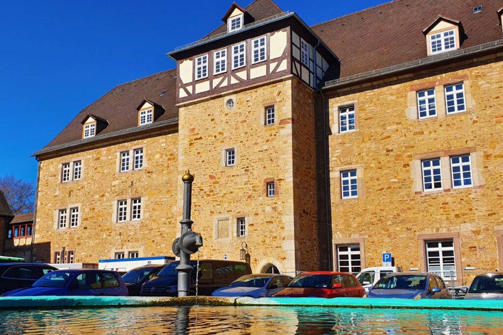 Das Melsunger Landgrafenschloss aus hellem Sandstein mit einem plätschernden Brunnen im Vordergrund und parkenden Autos auf dem Vorplatz.