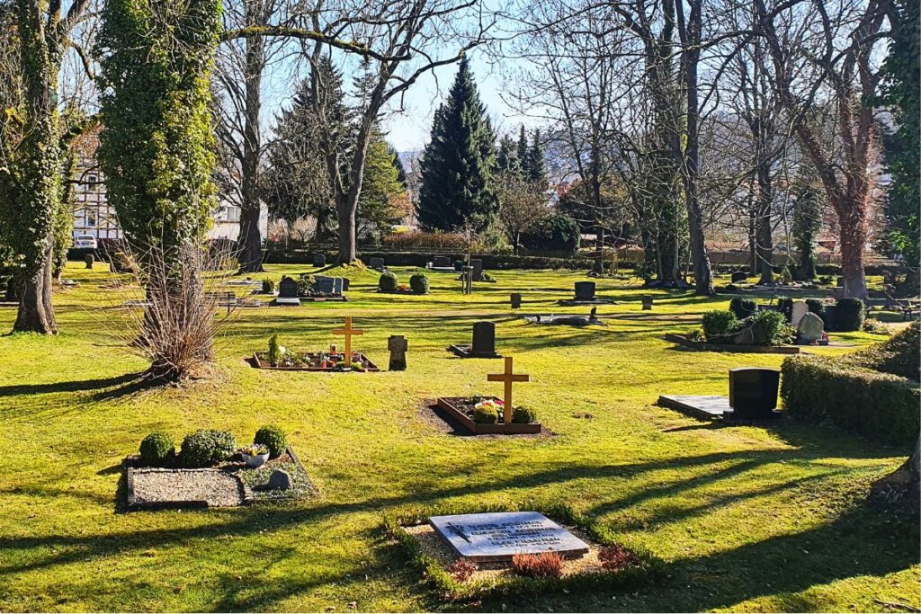 Blick über den Alten Friedhof in Melsungen mit grünen Rasenflächen, schattenspendenden Bäumen und historischen Grabkreuzen im Sonnenlicht.