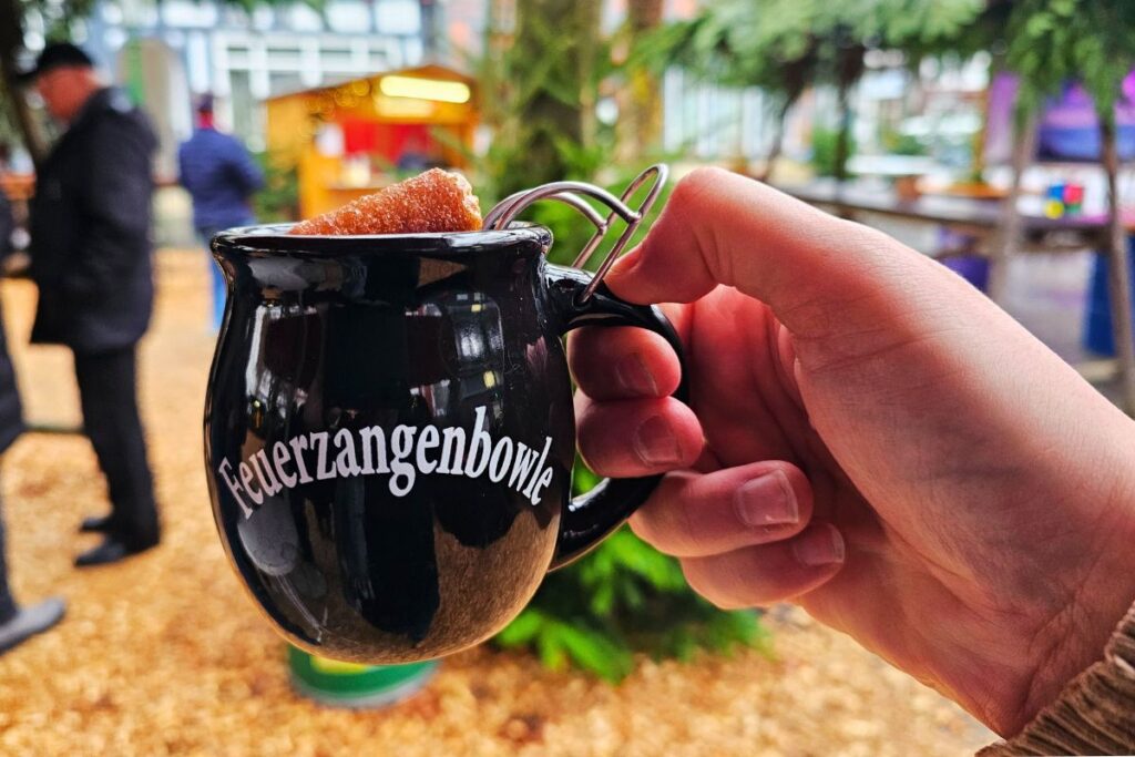 Eine Hand hält eine dunkle Tasse mit der weißen Aufschrift „Feuerzangenbowle“ auf dem stimmungsvollen Melsunger Weihnachtsmarkt.