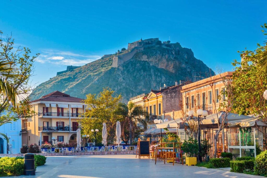 Blick über eine sonnige Straße in Nafplio mit neoklassizistischen Gebäuden auf die mächtige Palamidi-Festung, die auf einem steilen Hügel thront.