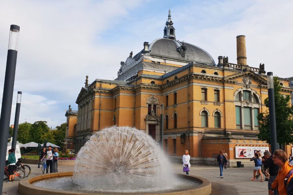 Das historische Nationaltheatret in Oslo aus gelbem Backstein mit grauer Kuppel, davor ein sprudelnder Kugelbrunnen