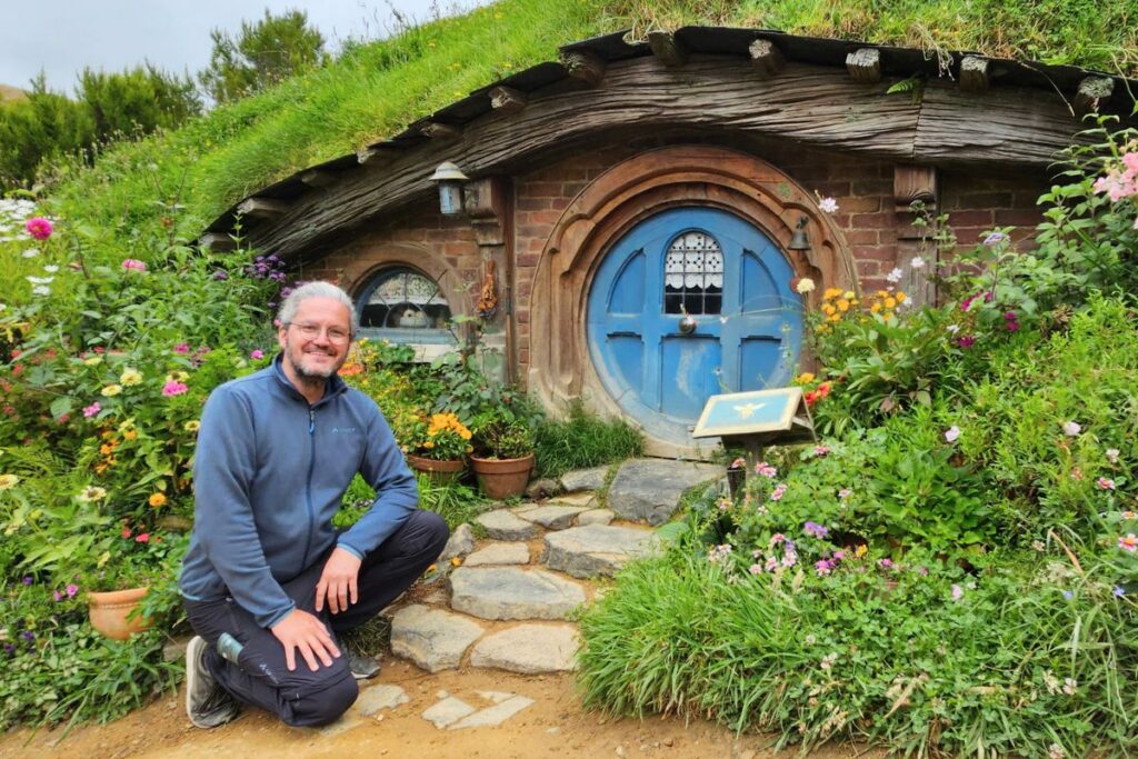 Reiseblogger Nicolo kniet lächelnd vor einer blauen Hobbithöhle mit runder Tür und üppigem Blumengarten im Hobbiton Movie Set.