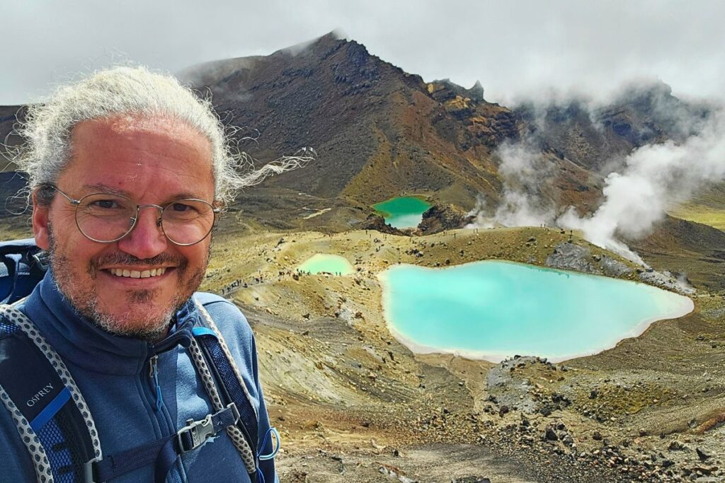 Reiseblogger Nicolo Martin lächelt vor den drei leuchtend türkisfarbenen Emerald Lakes und dampfenden Schwefelfeldern im Tongariro Nationalpark.